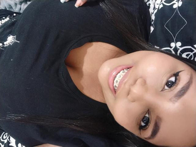 El rostro de Sarah Smity, con cabello castaño oscuro, una camisa negra y dientes sonriendo que muestran brackets metálicos. Tiene ojos marrones y un tono de piel ligeramente bronceado..