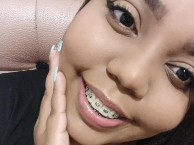 O rosto de Sarah Smity, sorrindo com aparelhos ortodônticos nos dentes, tem pele marrom clara, cílios longos e ela está tocando sua bochecha com uma mão que tem unhas azul claras..
