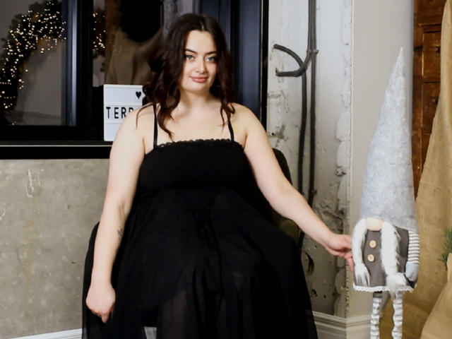 MilanaPrice with curly dark brown hair, wearing a black, sleeveless, strapless dress, holding a small, white, fuzzy, toy figure with brown buttons. Smiling gently.