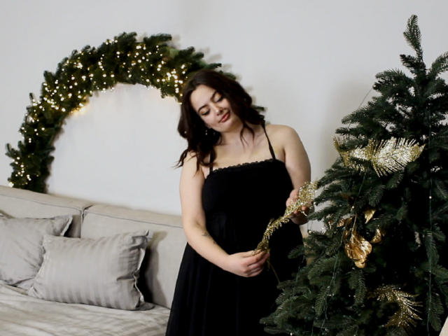MilanaPrice, com cabelo castanho ondulado, vestido preto e seios pequenos, decorando uma árvore de Natal com enfeites dourados, sorrindo, em pé em frente a uma guirlanda verde iluminada..