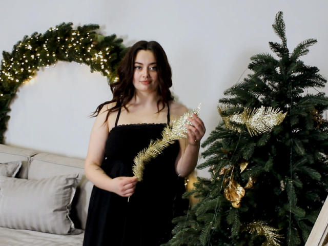 MilanaPrice è in piedi, con i capelli scuri e ondulati, indossa un abito nero, tiene in mano un festone dorato, di fronte a un albero di Natale decorato con ornamenti dorati e luci bianche..