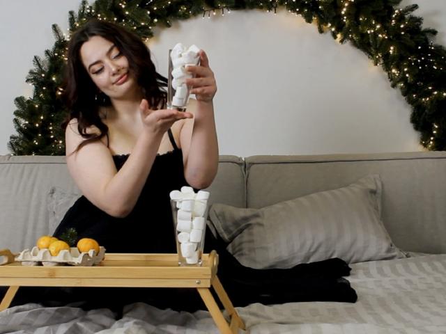MilanaPrice with medium tits, black dress, dark hair, holding marshmallows, pouring into a glass, on a gray couch, wooden tray with oranges, green garland with lights.
