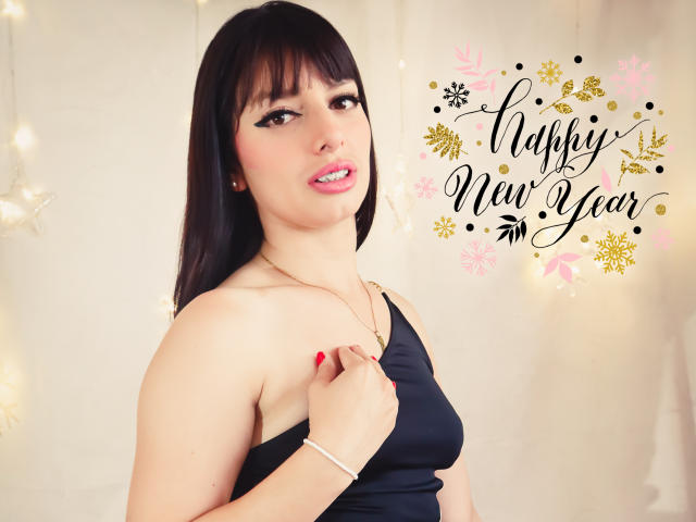 LeticiaFlorenz, con cabello castaño oscuro, un top de tirantes negros, uñas rojas, un collar dorado y una ligera sonrisa, sosteniendo su top, mostrando sus pequeños senos. Luz suave, fondo blanco..