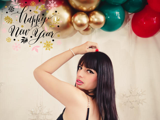 LeticiaFlorenz, com cabelo preto, olhando por cima do ombro, com a mão no cabelo, vestindo uma blusa preta, unhas vermelhas, pulseira dourada, balões verdes, vermelhos e dourados acima, sorrindo, fofa, sexy..