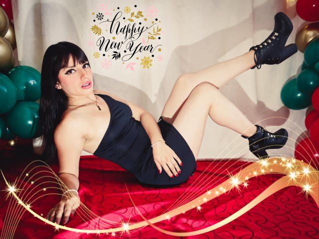 LeticiaFlorenz liegt auf einem roten Teppich, trägt ein schwarzes, trägerloses Kleid und schwarze High Heels mit Nieten, wobei ihre Brüste und ihr Hintern sichtbar sind, und hinter ihr befinden sich grüne Luftballons..