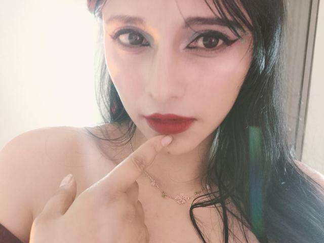 Midori Chang mit schwarzen Haaren, rotem Lippenstift und Eyeliner, die mit ihrem Finger ihre roten Lippen berührt, trägt eine rosa Blumenkette, hat helle Haut und einen verführerischen Blick..