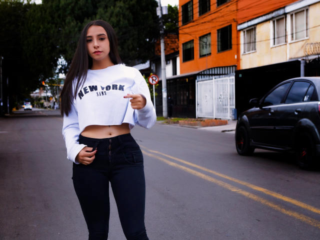 Kaiylin, met lang bruin haar, een witte crop top die haar platte buik laat zien, en strakke zwarte jeans, staat op een straat en wijst met haar rechterhand. Een zwarte auto op de achtergrond..