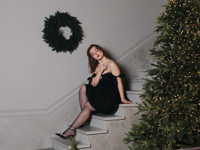 Stacy Yellig sentada nas escadas, vestindo um vestido preto, meias de rede e saltos, com as mãos no peito, ao lado da árvore de Natal, uma guirlanda verde na parede, com uma expressão ligeiramente entediada..