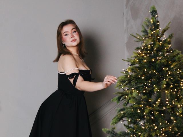 StacyYellig in a black dress, standing on stairs, touching a Christmas tree with lights, wearing fishnet stockings and high heels, looking back with a slightly open mouth.