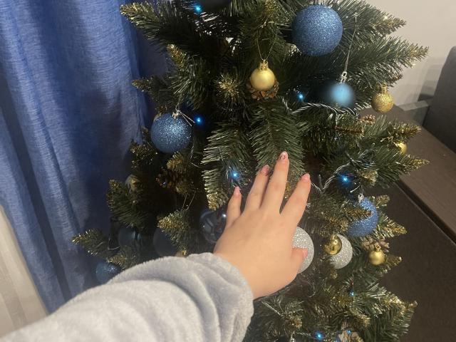 La mano de Dooris, con uñas rosas, toca un árbol de Navidad azul y dorado, adornado con adornos brillantes y pequeñas luces. Dooris lleva un suéter gris, suave y de manga larga..