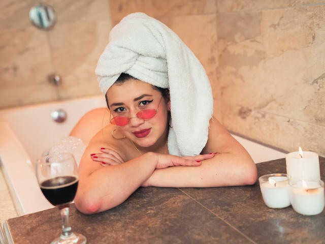 VeronicaHot69 avec une serviette blanche sur la tête, des lunettes roses, des ongles rouges et du rouge à lèvres rouge, penchée dans une baignoire, les bras sur le bord, un verre de vin, des bougies et du vin rouge..