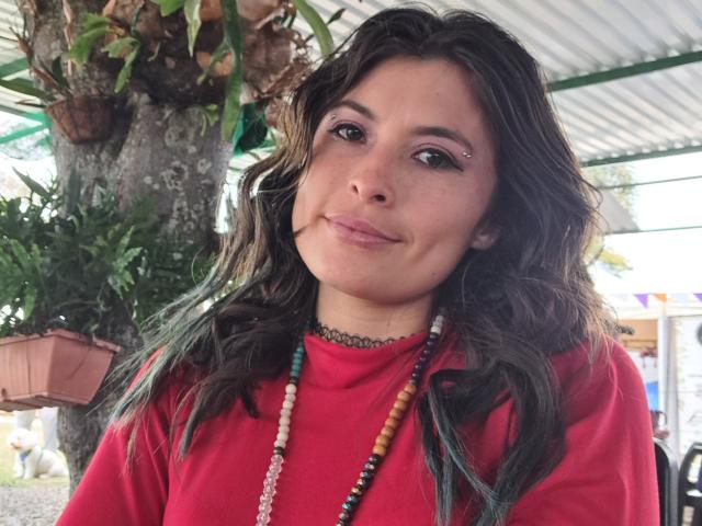 AlexandraSaenz69 heeft golvend donker haar, een rood shirt, een kralenketting en een neusring, ze glimlacht en heeft sproeten, draagt een zwarte choker, en staat onder een structuur met een groen dak..