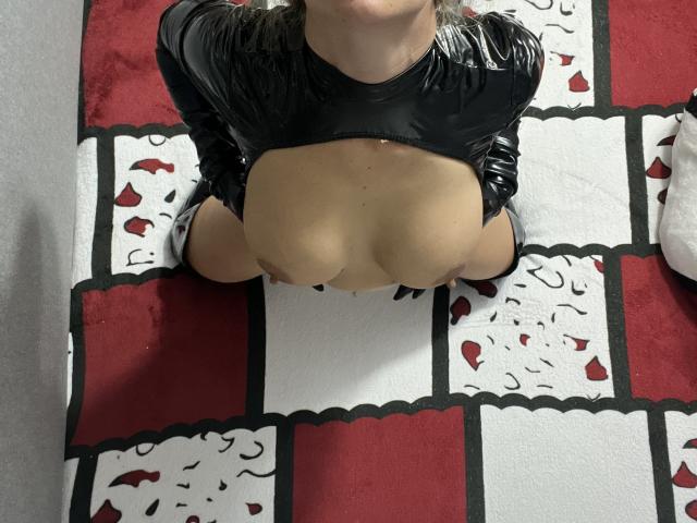Cyberxxx, gekleed in een zwart leren top, knielt met grote, onbedekte borsten, waarbij haar gebruinde huid zichtbaar is, en er ligt een rood-wit gestreept tapijt onder haar..