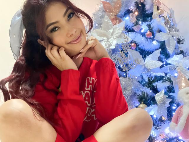 KendraMiler with red sweater, sitting in front of blue Christmas tree, smiling, hands on cheeks, long red hair, blue eyes, light skin, cute, sexy, Christmas decorations, white poinsettias.