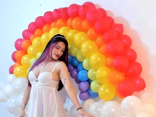 LeviGold with purple hair, white dress, and small tits, standing in front of a colorful balloon arch. She's smiling, arms out, wearing a white headband.