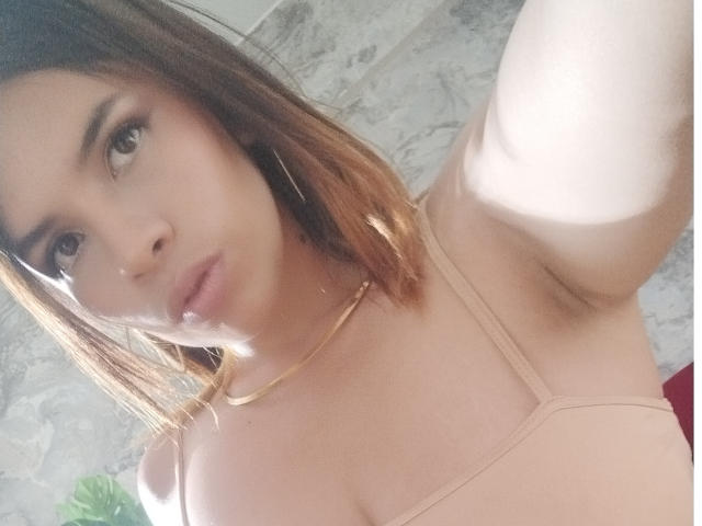 VioletaStone with light brown hair, wearing a beige top, showing her tits, armpit, and pouty lips, in a selfie with a gold necklace and hoop earrings.