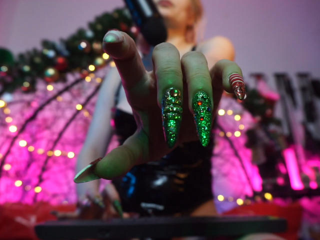 De hand van CyberXGoddess, met lange, glinsterende groene nagels en rode stipjes, wijst vooruit. Ze draagt een zwart masker en een zwart latex outfit, met roze en paarse verlichting..