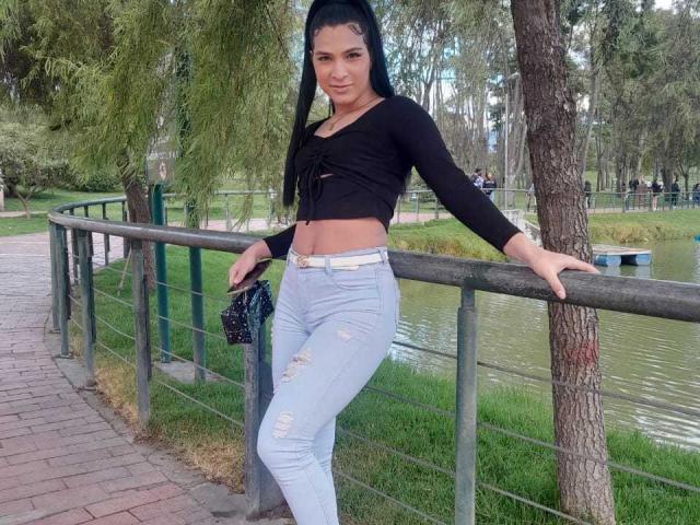 BarbaraParker with long black hair, black crop top showing her tits, light blue ripped jeans, standing against a fence, holding a black purse, in a park.