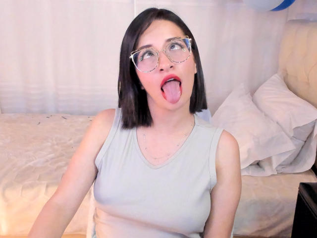 LeviGold, con cabello negro corto, gafas y una camiseta gris claro, saca la lengua, mostrando una expresión juguetona y sorprendida. Sus pequeños senos son ligeramente visibles..