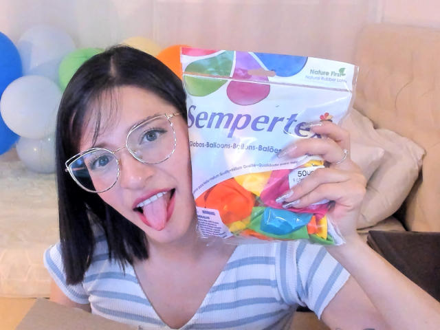 LeviGold with black hair, glasses, and tongue out, holding a Semperit condom package, wearing a striped shirt, sitting on a beige couch with balloons in the background.
