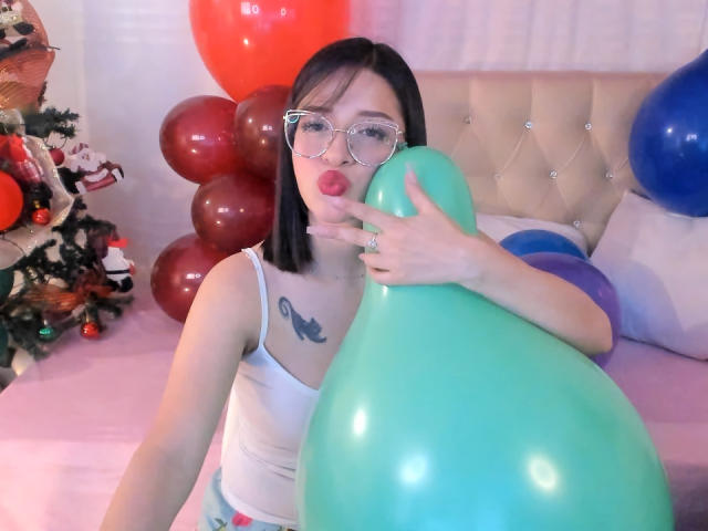 LeviGold con un corte de pelo bob negro, gafas, camiseta blanca, sosteniendo un globo verde, labios rojos y gruesos, tatuaje en el pecho, cama rosa, globos rojos y azules, árbol de Navidad a la izquierda..