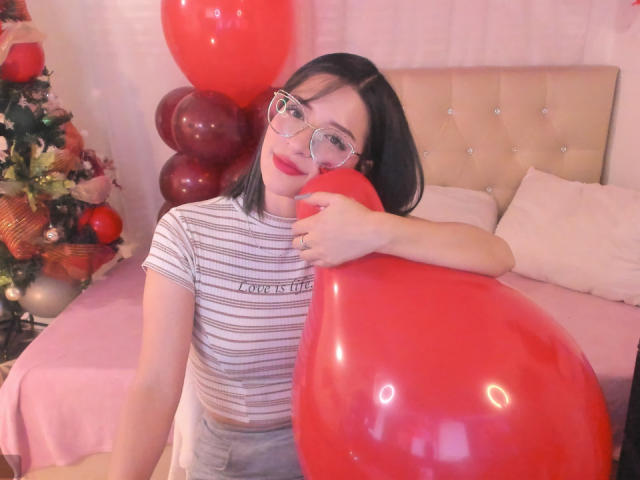 LeviGold, avec des lunettes, cheveux noirs courts, un ballon rouge, une chemise rayée, et un lit rose, tenant un ballon rouge, à côté d'un sapin de Noël avec des décorations rouges..