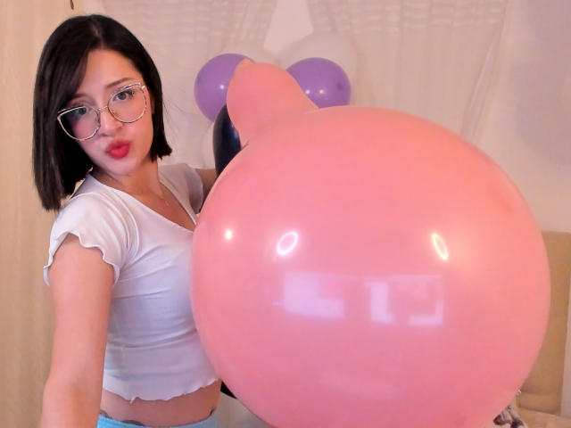 LeviGold mit kurzen, schwarzen Haaren, Brille und roten Lippen, hält einen riesigen, rosa Luftballon, trägt ein weißes Crop-Top, das ihre kleinen Brüste zeigt, und hat ein süßes Gesicht..