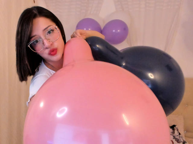 LeviGold con occhiali, rossetto rosso e taglio di capelli corto nero, che tiene palloncini rosa e blu, concentrandosi sul grande palloncino rosa davanti a lei. Il seno, i glutei e i palloncini sono ben visibili..