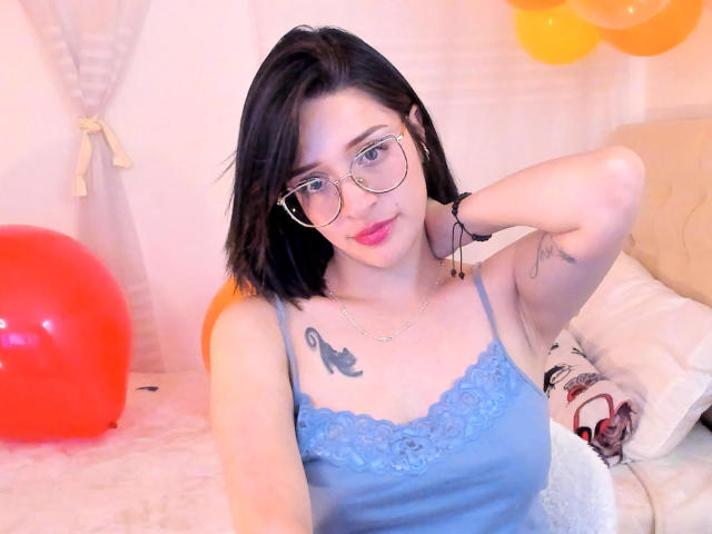 LeviGold mit schwarzen Haaren, Brille und einem blauen Spitzenoberteil, zeigt ein Vogel-Tattoo auf ihrer Brust, lächelnd, Hand hinter dem Kopf, orange und gelbe Luftballons, roter Ball..