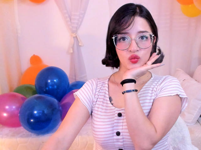 LeviGold with short black hair, glasses, pouty lips, white striped shirt, black button, blue balloon cluster, right hand on cheek, black wristbands, soft pink and orange balloon backdrop.