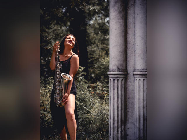 GinaONeon with brown hair, black dress, holding a silver saxophone, smiling, standing between white marble columns, sunlight on her ass, green foliage in background.