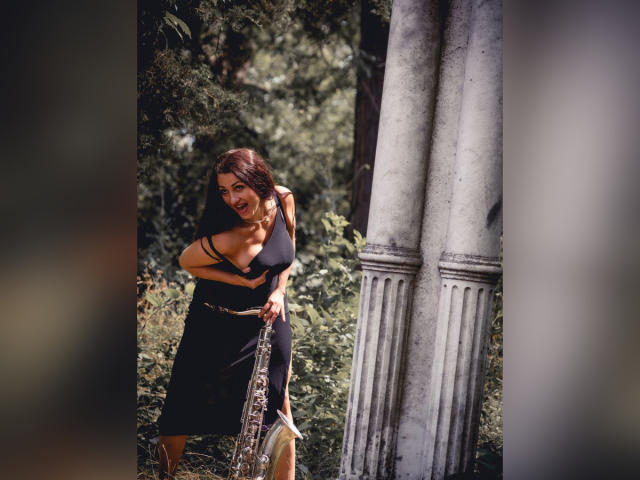 GinaONeon with brown hair, laughing, wearing a black dress, holding a silver trumpet, standing next to white, fluted columns, in a sunny, green, wooded area.