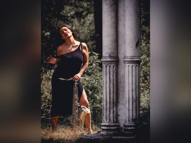 GinaONeon stands in a sun-dappled ruin, wearing a tight black dress with a thigh slit, holding a broken column, showing her toned arms and legs, with her ass visible.