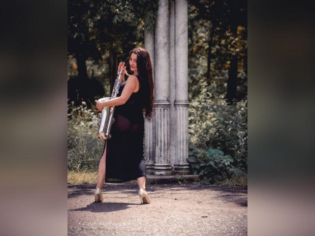 GinaONeon, com cabelos longos e escuros, vestindo um vestido preto sem mangas e saltos altos, segurando uma trompete prateada, em pé em frente a uma coluna de mármore, exibindo suas nádegas, em um parque..