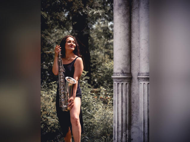 GinaONeon, com cabelos castanhos longos, vestindo uma camiseta preta e shorts, segurando um saxofone prateado, em pé perto de colunas brancas, sorrindo com os olhos fechados..