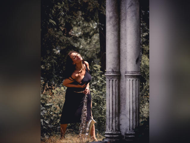 GinaONeon se tient debout, les bras croisés, vêtue d'une robe noire, avec des cheveux bruns ondulés, appuyée contre un pilier, la lumière du soleil sur ses seins, une forêt verdoyante, et une chaîne dans sa main droite..