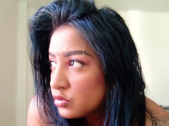 Nahirr has dark, wet-looking black hair, brown eyes, and a nose ring. She has a small bindi on her forehead and a subtle lip piercing. She looks to the side.