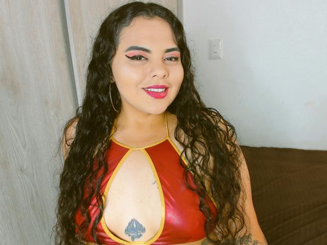 ElizaBliss, con cabello negro largo y rizado, lleva puesto un top de cuero rojo con tirantes, que revela sus pechos y un pequeño tatuaje debajo. Está sonriendo y lleva pintalabios rosa..
