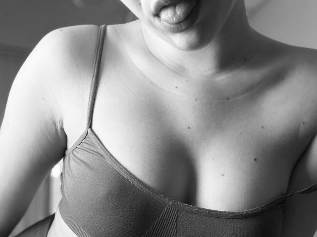 SugarrX with tongue out, wearing a sports bra, showing her tits and a few small moles on her chest. Black-and-white photo, upper body focus, slightly blurred.