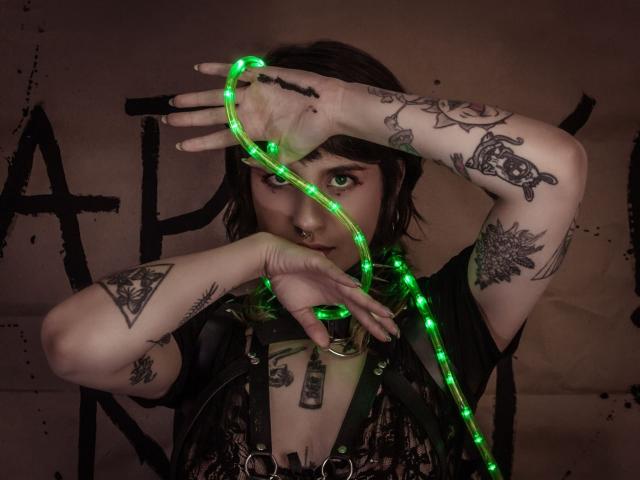 AmySpider mit grünem Neonkabel über dem Gesicht, schwarzem Ledergeschirr, Tattoos, Nasenring und kurzen, dunklen Haaren, die ein Neonkabel in der Nähe ihrer Brüste hält. Dunkles, kantiges, punk-inspiriertes Aussehen..
