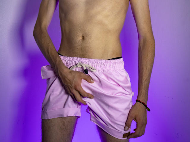 O corpo esguio e tonificado de DamianMaya, com costelas visíveis, vestindo shorts rosa, com a mão segurando a parte da frente, puxando-os levemente para baixo, revelando um pouco de roupa íntima preta, fundo roxo claro..