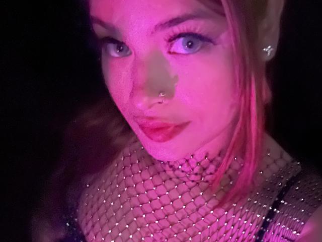 Shantal, met roze haar, blauwe ogen, een neusring en rode lippenstift, draagt een glinsterende top van gaas en kijkt met een lichte glimlach in de camera. Roze en paarse verlichting..