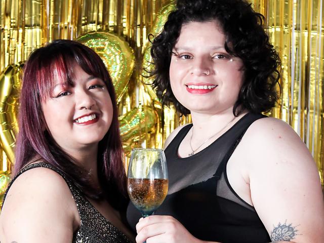 IsaAndCaro mit roten und schwarzen Locken, lächelnd, mit einem Glas Bier in der Hand, in schwarzen Kleidern, goldene Ballons im Hintergrund, ihre Brüste sind leicht unter dem Kleid sichtbar..