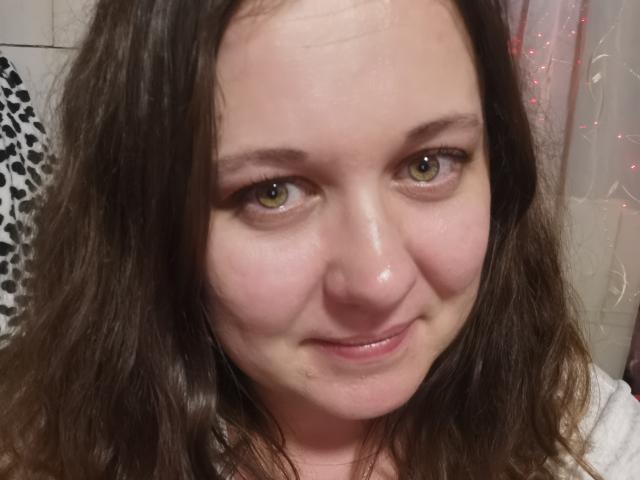 ChaudeBella69 heeft groene ogen, golvend bruin haar en een lichte glimlach met een klein beauty markje op haar kin. Haar huid is licht en ze draagt een witte top..