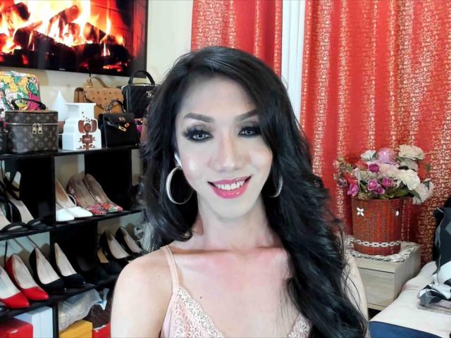 Una mujer llamada OneFoxyDiva, con cabello negro largo, vistiendo una blusa rosa de encaje, aros grandes y lápiz labial rojo, sonriendo en una habitación con cortinas rojas y estantes para zapatos. Se ven los senos..