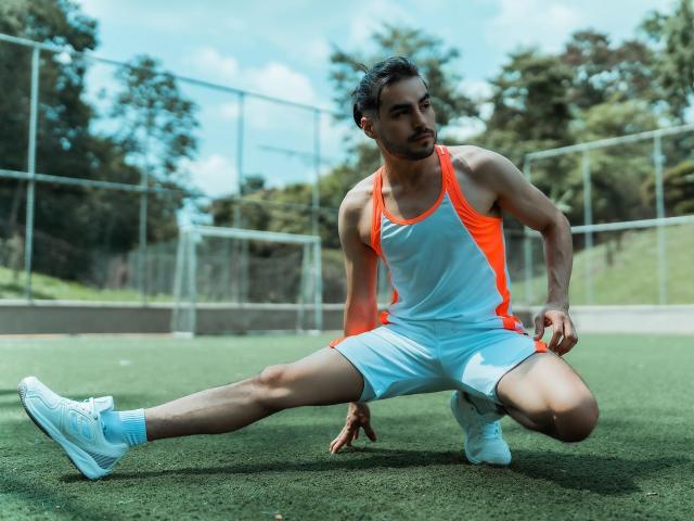 MateoGrand, musculoso, com pele escura e cabelo preto curto, ajoelha-se na grama verde, vestindo uma regata esportiva branca e laranja, demonstrando um alongamento enorme e forte da perna, usando tênis brancos..