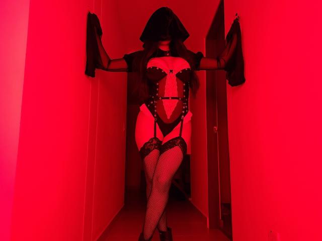 MeganWisse in red-lit hallway, wearing black hood, sheer mask, black lace corset, garter belt, fishnet stockings, and heels. Hands on walls, legs crossed, exposing her pussy.
