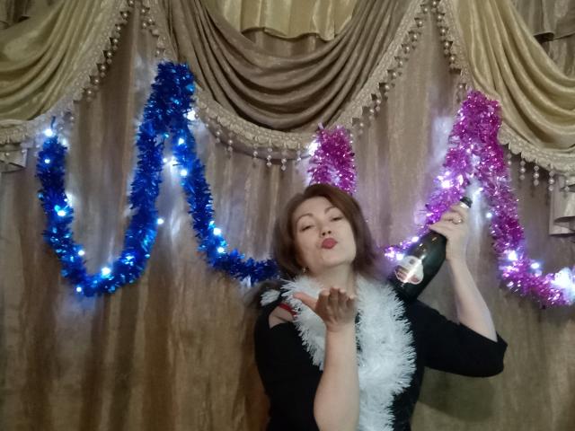 SadieLorri, con cabello castaño, labios fruncidos, una blusa negra, una pluma blanca, sosteniendo una botella, adornos de tinsel azules y rosas, luces, frente a cortinas con drapeado. Los senos están cubiertos por la blusa..