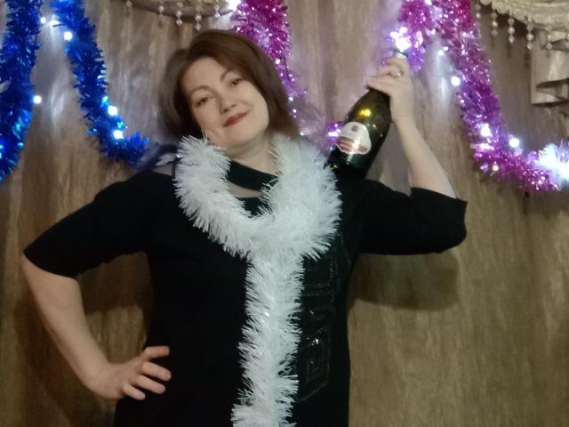 SadieLorri, com cabelo castanho, vestido preto, guirlanda branca, segurando uma garrafa, sorrindo, com guirlanda azul e roxa ao fundo, com a mão na cintura..