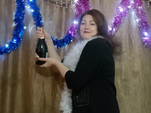 SadieLorri with brown hair, black top, white feather boa, holding a bottle, smiling, blue and pink tinsel, and lights in the background. Her tits are covered by the top.